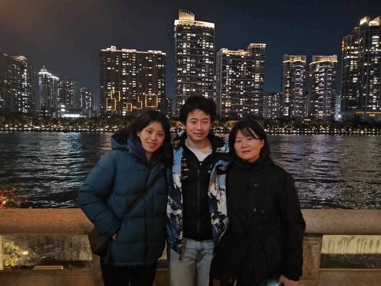 Three people stand in front of a water body with brightly lit tall apartment buildings behind them.