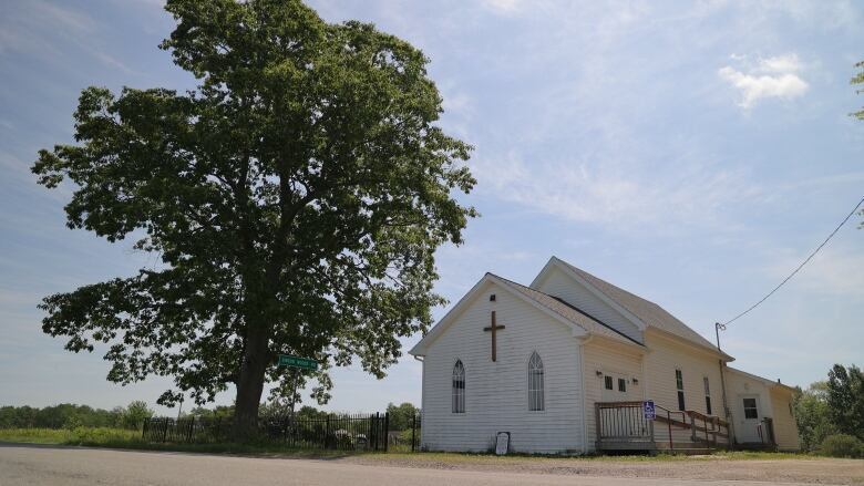 A small white church on a rural road, next to a large tree, under a blue sky.