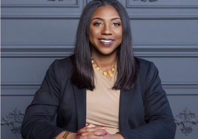 A woman in a blazer sits at a desk and smiles into the camera. 