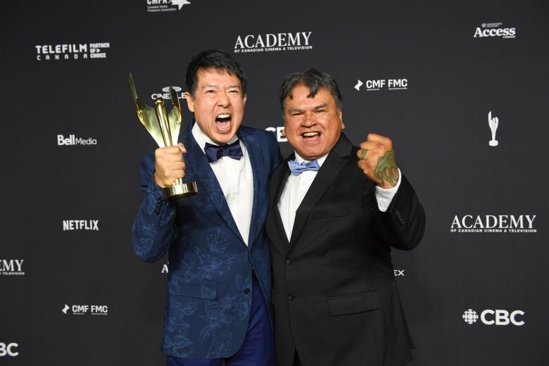 Two men in tuxedo punching their fist in victory, while one of them holds an award.