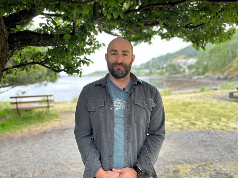 A man with a beard, wearing a grey jean jacket, stands in front of a body of water.