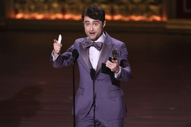 A person in formal wear gestures while making a speech and holding an award.