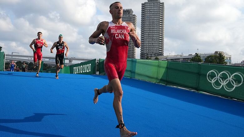 A Canadian men's triathlete, dressed in a red outfit with white lettering, runs on a blue track with a banner reading 