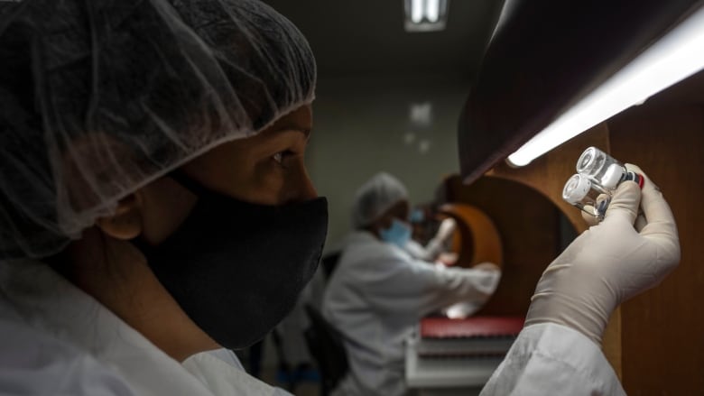 A scientist in a white haircap and gloves holds up a vial.