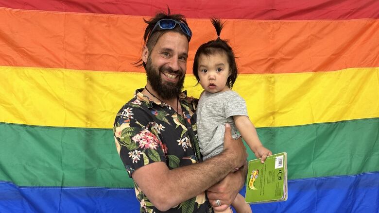 A man stands in front of a Pride flag, holding a small child and smiling.