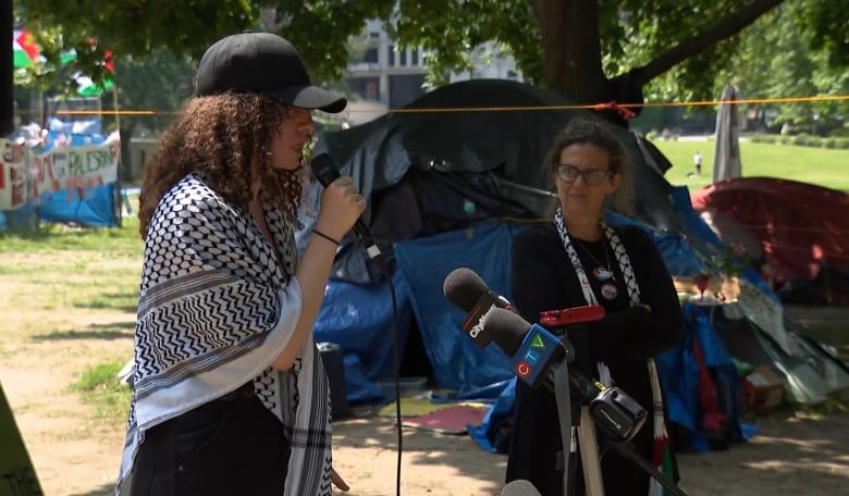 A young woman with a cap and keffiyeh scarf speaking into a microphone 