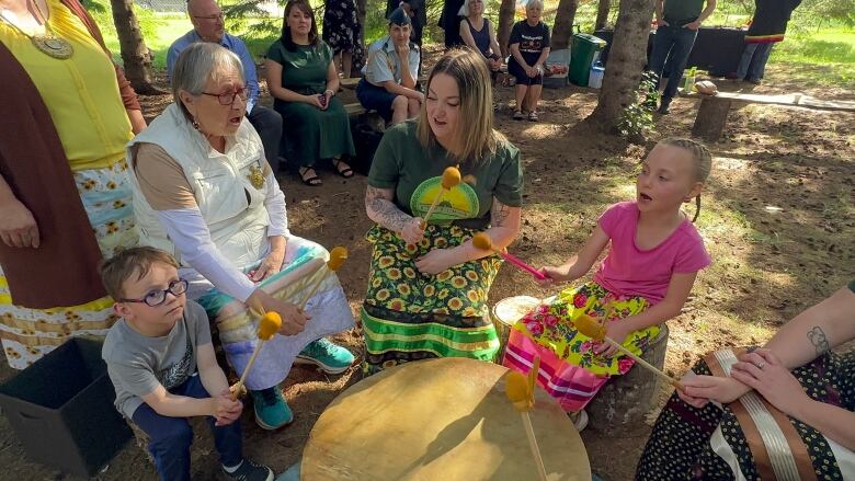 A group of adults and children sit around a drum in a forest. They hold drum sticks in their hands.