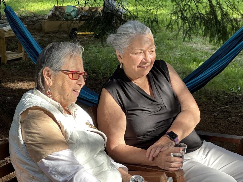 Two women with grey hair sit together outside in a forest. A blue hammock stretches behind them.