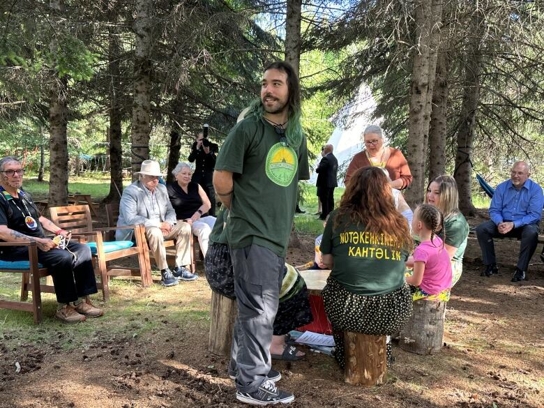 A man wearing a green t-shirt with black and green hair stands in front of a group of people sitting in a circle in a forest.