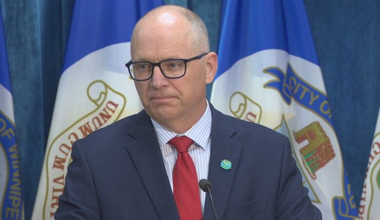 A bald man with glasses wears a blue suit and stands at a podium.