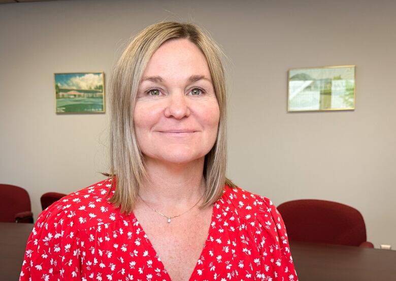 A woman with blond hair wearing a red shirt with a white print pattern sits in a boardroom and smiles.