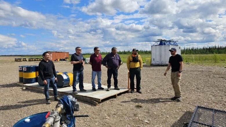 A line of people listen to another person while standing on gravel.