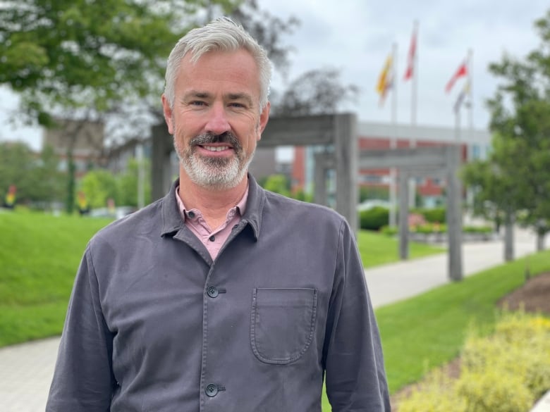 A man with short grey hair and a grey beard, wearing a button down shirt over a pink t-shirt, is pictured outdoors at the University of Waterloo.