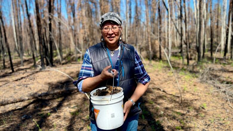 a woman in blue jeans walks in a forest and carries a white bucket full of morel mushrooms