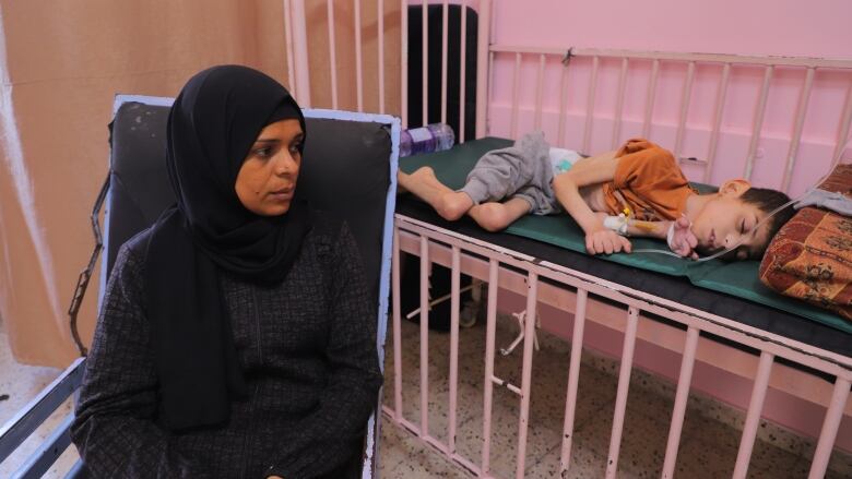 A woman in black sits on a black chair. Behind her is an emaciated child lying in bed. They are in a hospital. 