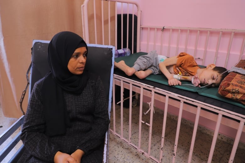 A woman in black sits on a black chair. Behind her is an emaciated child lying in bed. They are in a hospital. 