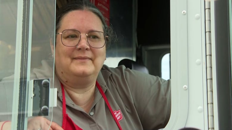 A woman with large framed glasses wearing an apron sits in the passenger seat of a food truck.