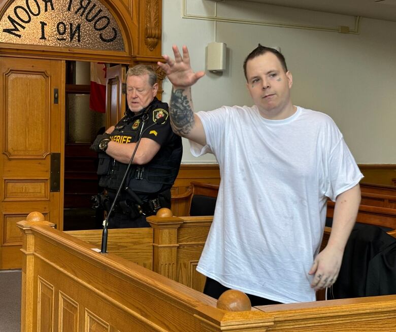 A man in a white t-shirt waving while standing in the prisoner's box of a courtroom.