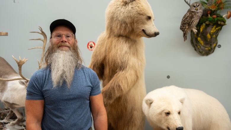 A white man in a blue shirt with a big beard and glasses. He's standing in front of two stuffed bears.