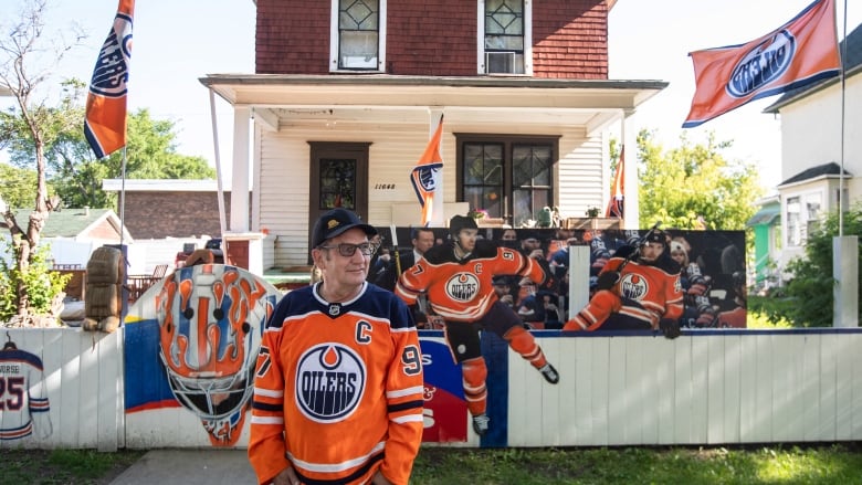 A man in an Orange and blue Oilers jersey stands in front of a house with Oilers flags and paintings of players.