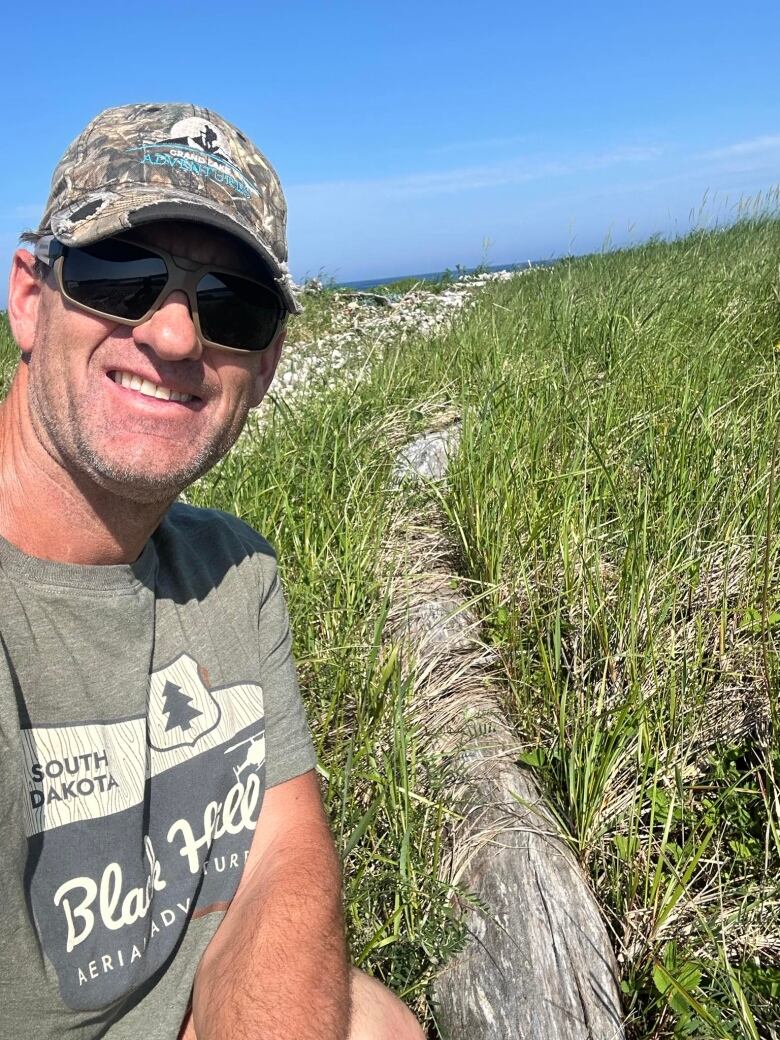 A man wearing a ball cap, sunglasses, and a sage green t-shirt is crouched in grass near a grey, weathered log.