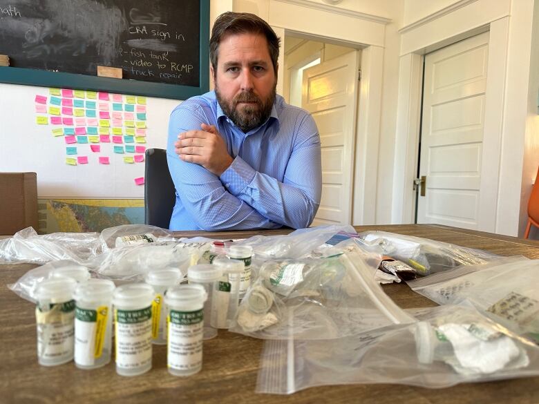 A white man wearing a blue shirt looks at a table with pill bottles and plastic bags on it.