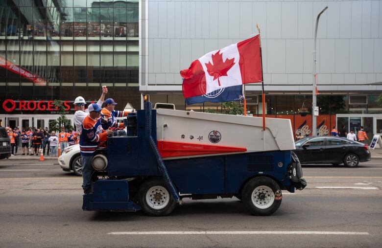 three men stand on a Zamboni driving on the street with a Canadian flag flying on it. 