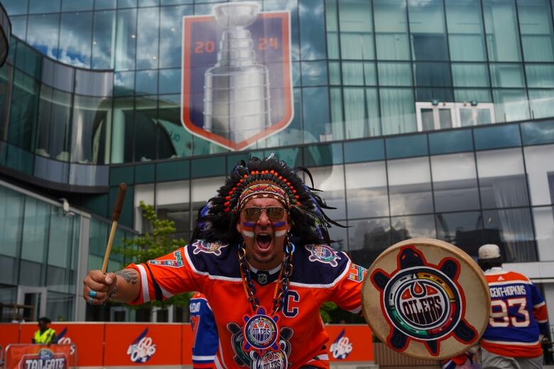 A man with red face paint, a headdress and a drum written Oilers appears to yell. 