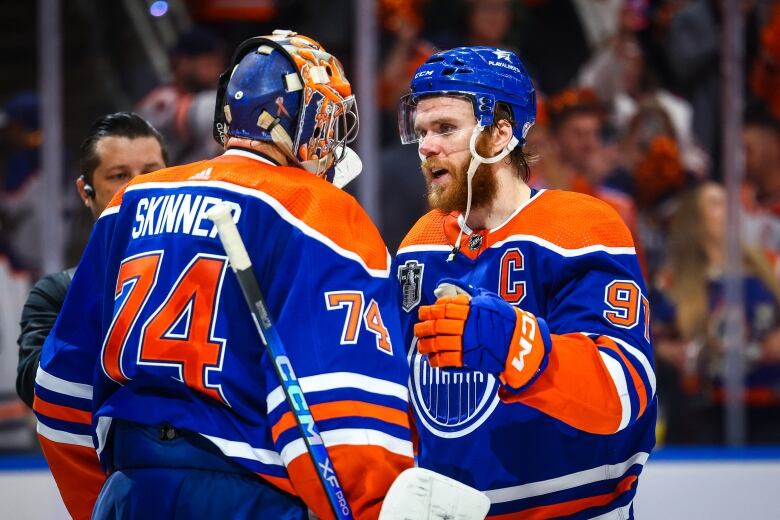 A male ice hockey player wrap his arms around his goaltender in celebration on the ice after a game as fans watch from behind the glass.