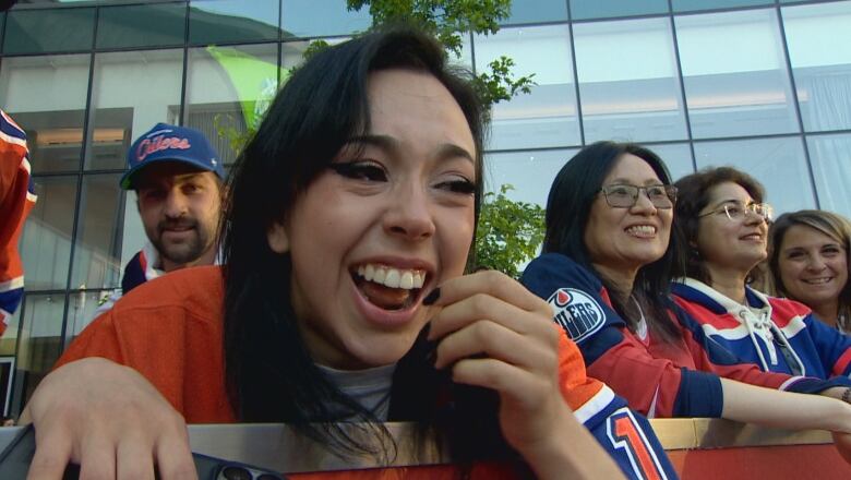 A woman with light skin and long black hair is smiling, with tears in her eyes, while wearing a blue-and-orange hockey jersey. She is outside, with other people around her smiling.