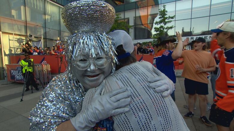 A woman in a silver costume and silver face paint is hugging someone outside, amongst a crowd of people.