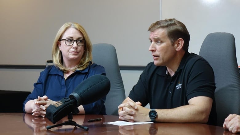 A man and woman sit at a table speaking into microphones