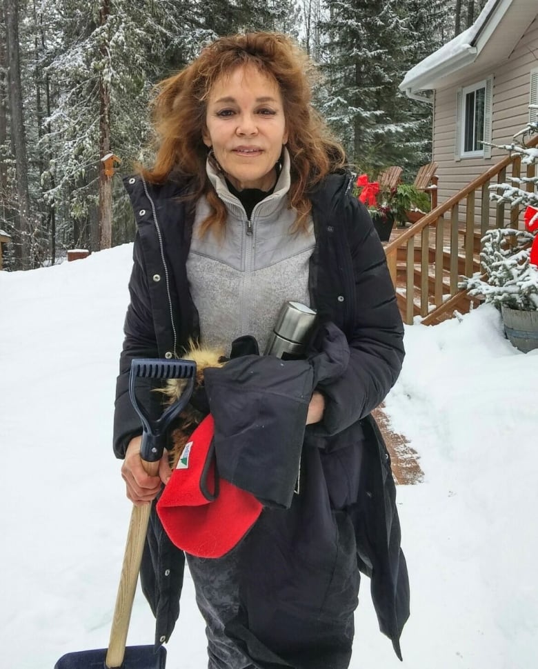 A woman with her winter jacket open holds a shovel in her hands. The deck behind her is free of snow. 