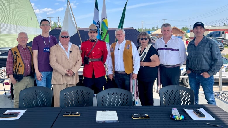 Eight people of varying heights and ages, including one woman, stand side-by-side on an outdoor platform, behind a table where they signed an agreement