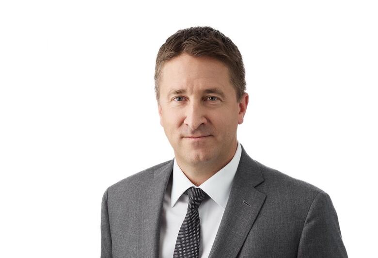 A man in a grey suit and tie is standing against a white background.