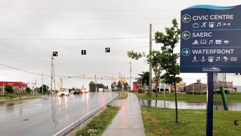 A sign in the foreground points to different buildings in different directions, while three arrow signs point different directions overhead of four lanes on a rainy street.