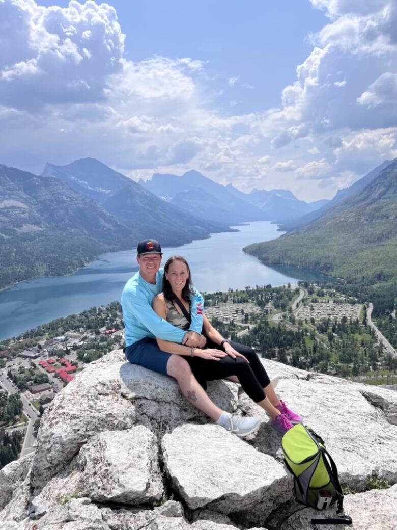 A couple sit on a rock with the mountains and a river flowing behind them.