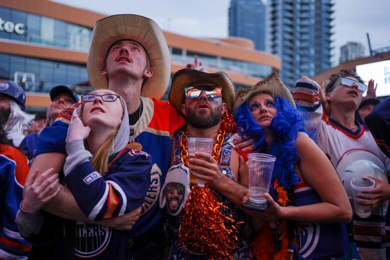 A group of people in Oilers gear stare upward at a screen 