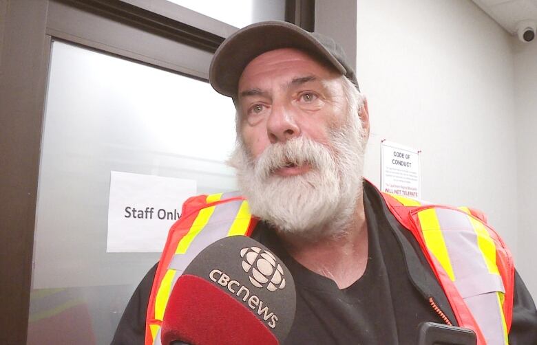 A man wearing a fluorescent orange and yellow vest and a black shirt with a grey ball cap and a big white beard and moustache speaks into a microphone.