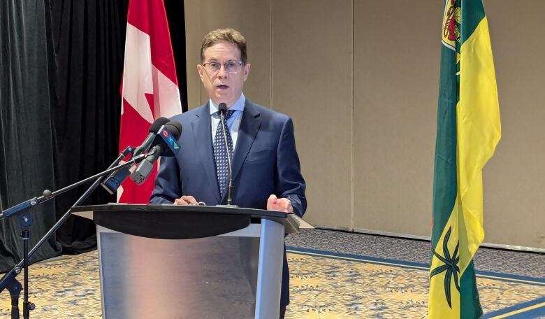 A man stands at a podium with Canadian and Saskatchewan flags behind him