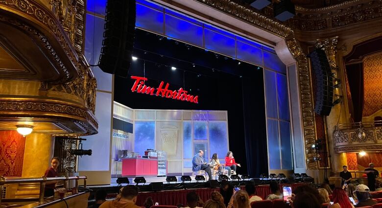 Three people sit on a stage in front of an audience. Overhead is a banner with the words Tim Hortons.