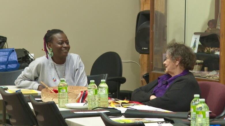 Fadeke Agboola sits at a table in a grey sweatshirt smiling at  Georgina Knockwood Crane who is in a black jacket. Green water bottles are on the table.