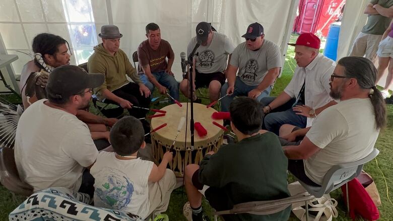 A group of men sit inside a white tent around a drum, each beating it with a stick and singing.