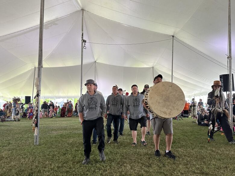 A group of men wearing grey sweaters walk through a white tent over grass. A man at the front holds a giant drum.