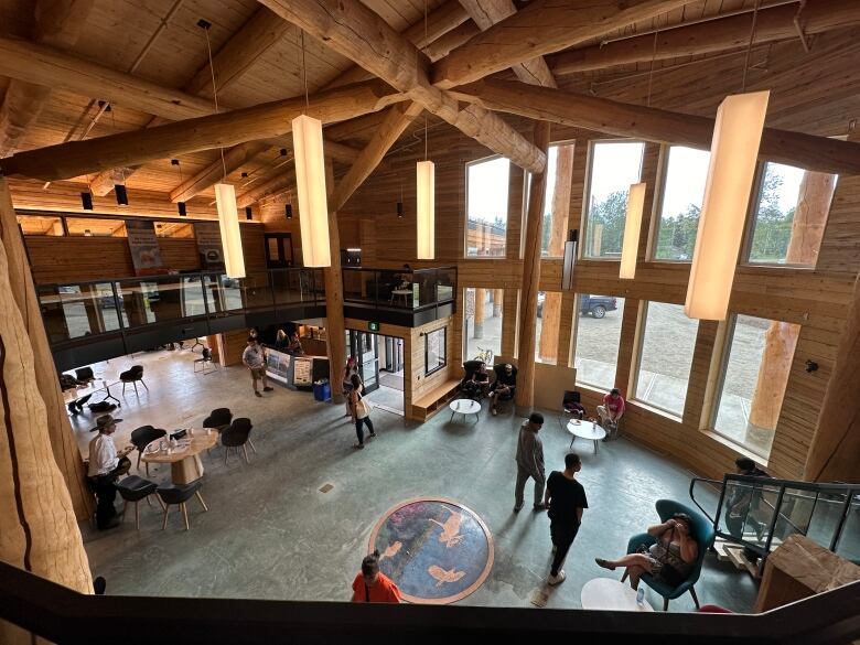 The interior of a light wooden building with large glass windows. A few people walk across the green floor below. 