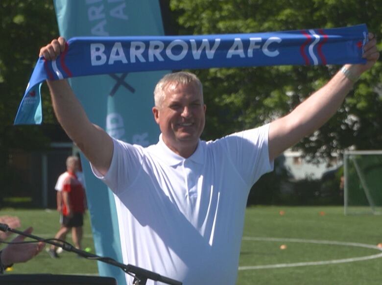 A man wearing a white t-shirt holds a blue scarf that reads, 'Barrow AFC.'