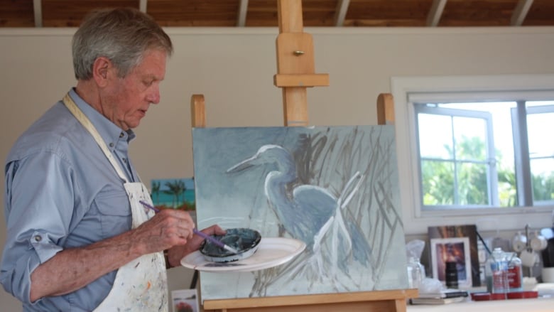 A man stands in front of an easel, holding a paintbrush. 