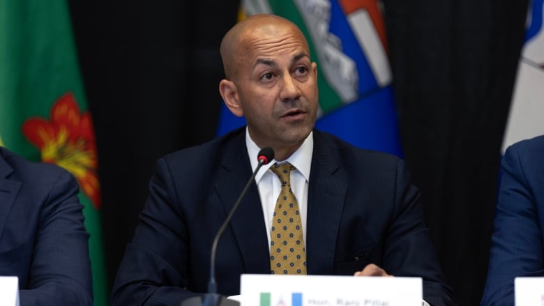 A bald man in a suit sits at a table speaking into a microphone.