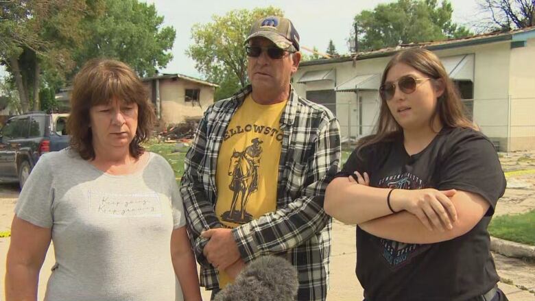 Three people stand together in front of a destroyed house.