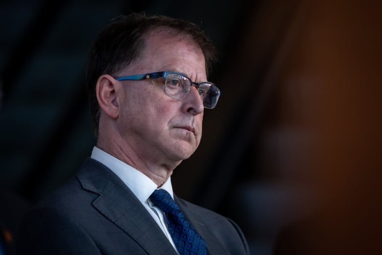 B.C. Health Minister Adrian Dix looks pensive at a press conference. He is a middle-aged white man wearing a navy blue suit, navy blue tie and white dress shirt. He has brown hair and wears black-framed glasses.